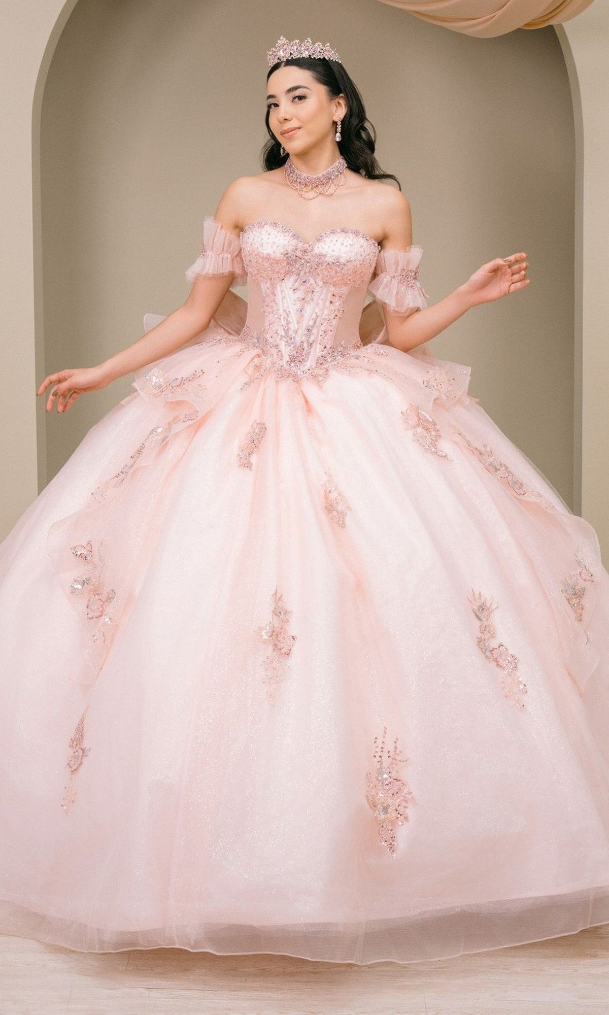 A woman wearing the Quinceanera Dress 8540JQ by Cinderella Couture in blush, an off-the-shoulder ball gown adorned with floral embellishments, stands in front of a neutral arch background, accessorized with a tiara and necklace.